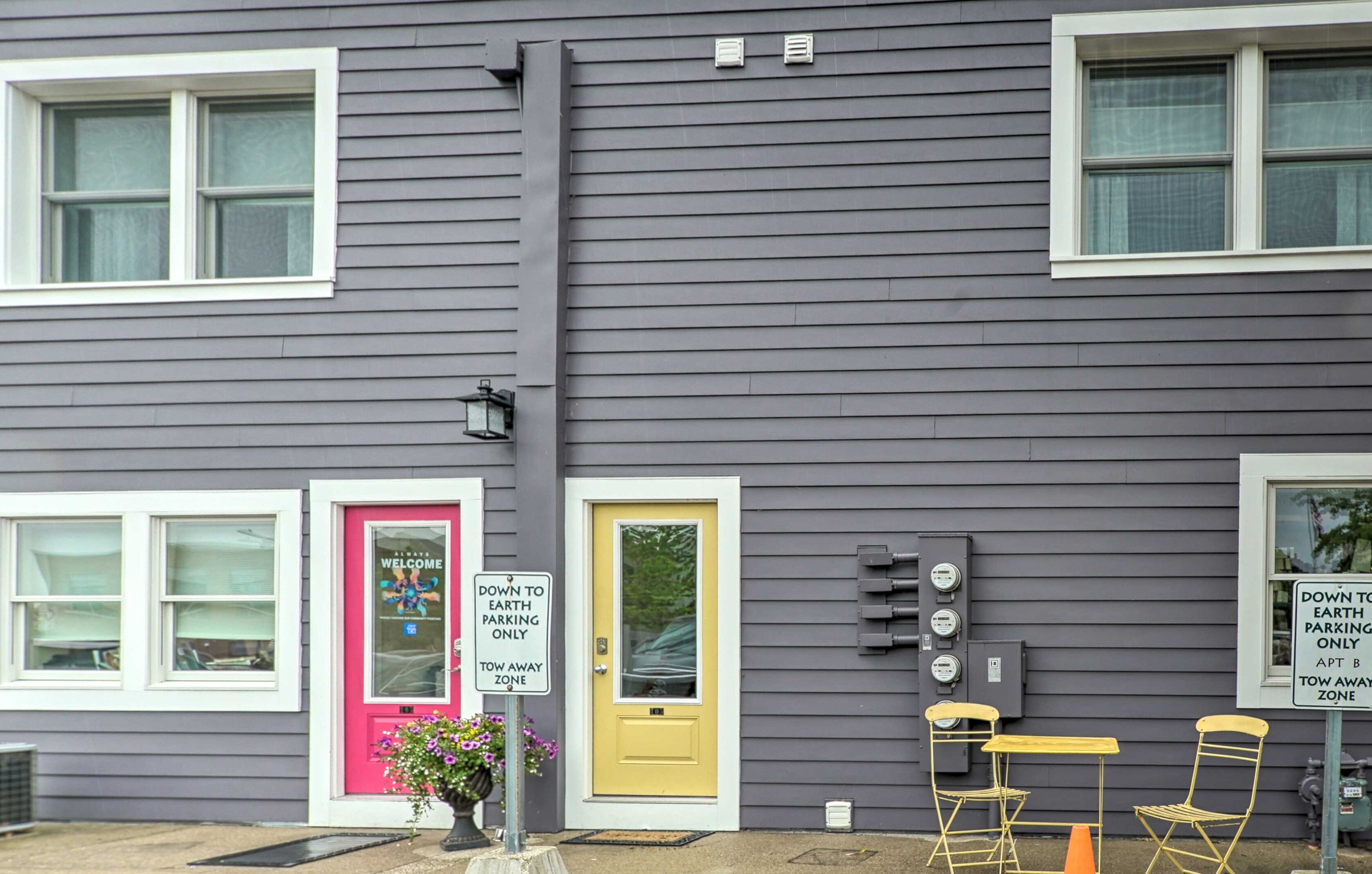 A building with a grey exterior, a yellow door, and a pink door with a 'Welcome' sign. There are two signs indicating 'Earth Parking Only' and a 'Tow Away Zone'.
