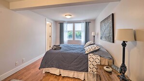 A bedroom with a bed, a bedside table with a lamp, a window with curtains, and a painting on the wall. - Cute Grand Haven Apartment: Walk Downtown Streets! (Grand Haven)
