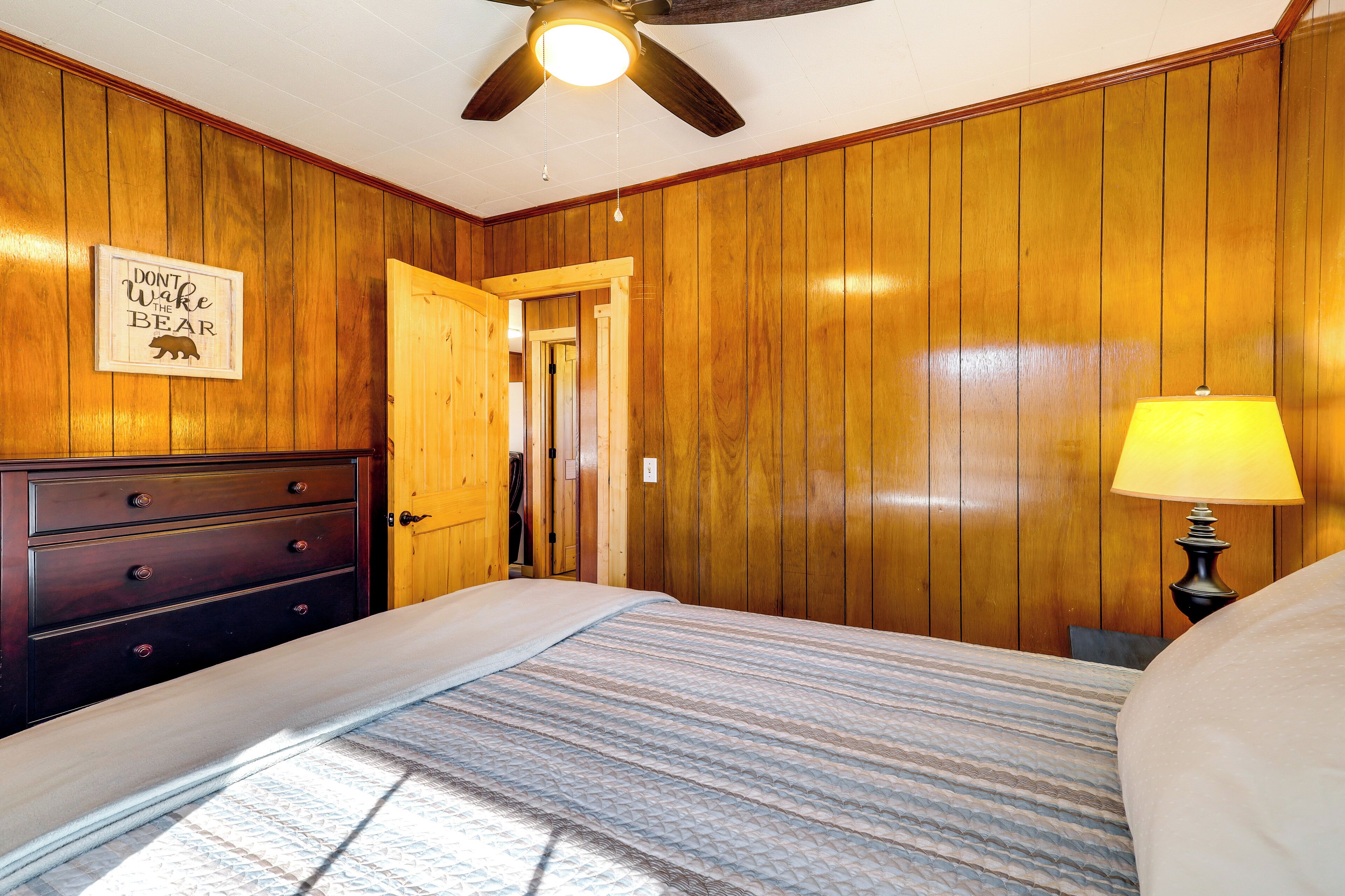 A bedroom with wooden paneling, a bed with a striped bedspread, a nightstand with a lamp, and a 'Don't Wake the Bear' sign on the wall.