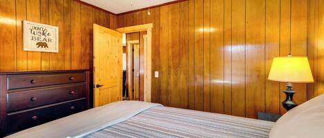 A bedroom with wooden paneling, a bed with a striped bedspread, a nightstand with a lamp, and a 'Don't Wake the Bear' sign on the wall.