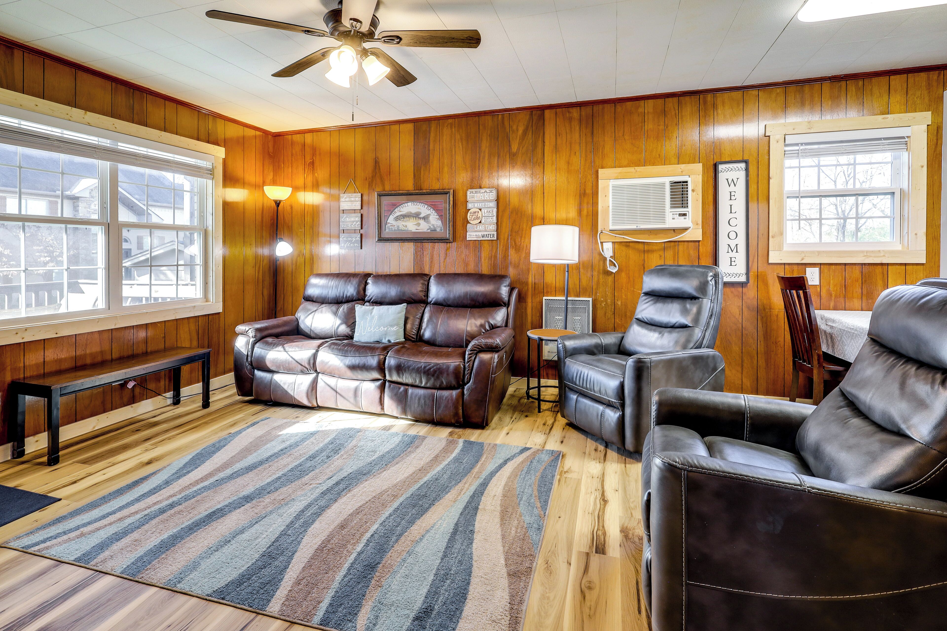 A cozy living room with wooden paneling, a ceiling fan, a brown leather sofa, a matching armchair, a striped rug, and a 'WELCOME' sign.