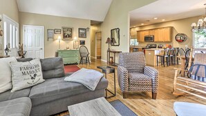 Un salón amplio con un sofá gris, un sillón estampado y un comedor con sillas de madera, con una cocina al fondo.
