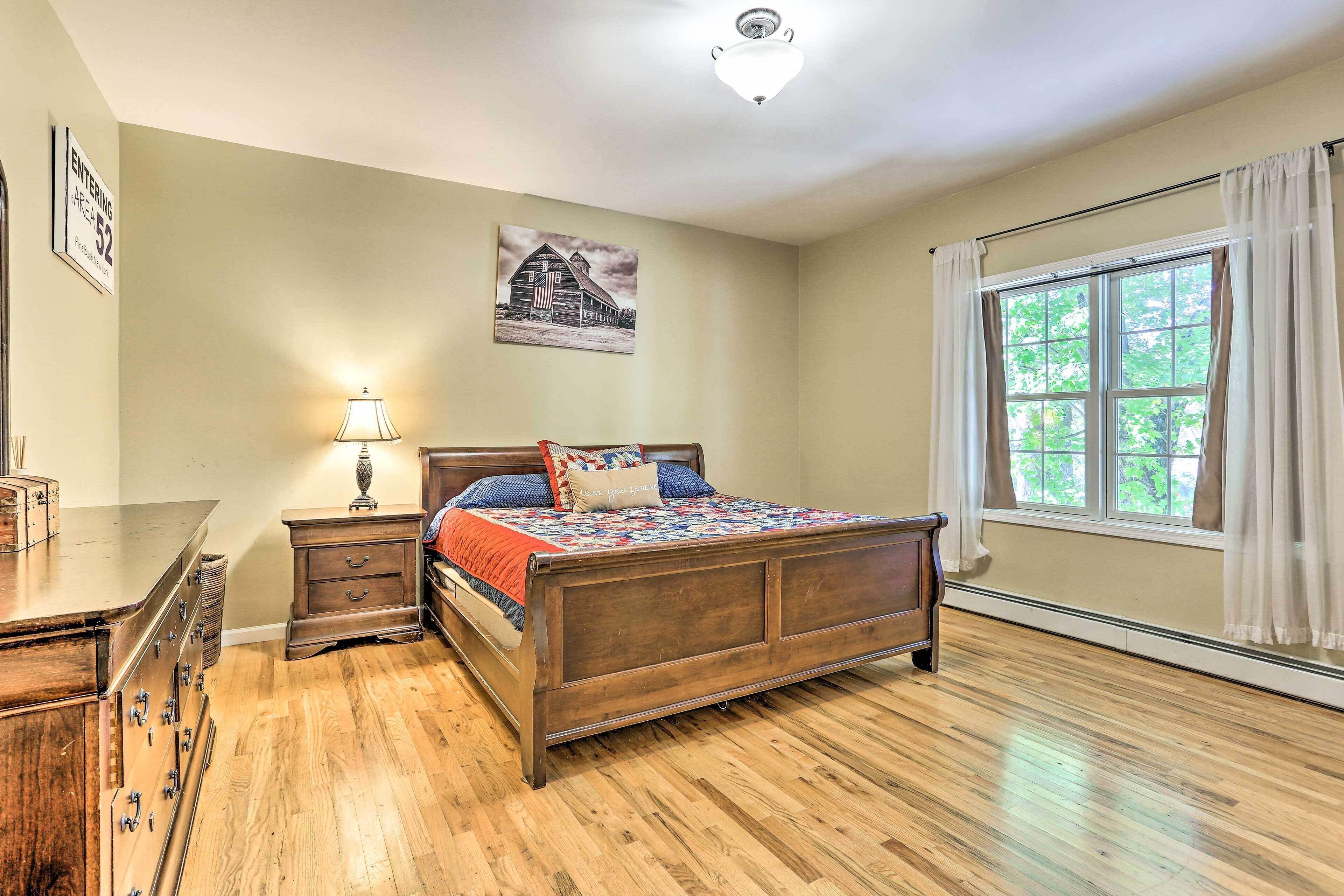 A bedroom with a wooden bed, a nightstand, a dresser, a lamp, a window with curtains, and a wall-mounted picture.