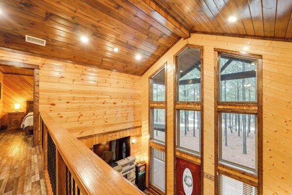 A wooden cabin interior with a fireplace, large windows, and a wooden staircase. - Broken Bow Cabin on 1 Acre w/ Hot Tub & Fire Pit! (Broken Bow)