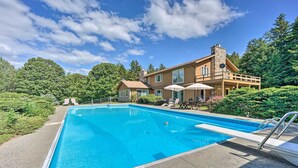 A rectangular swimming pool with a clear blue water, surrounded by a paved deck. There are lounge chairs and umbrellas near the pool, and a large house with a stone chimney in the background. - Family Winter Retreat w/ Hot Tub: 14 Mi to Slopes! (Whitingham)