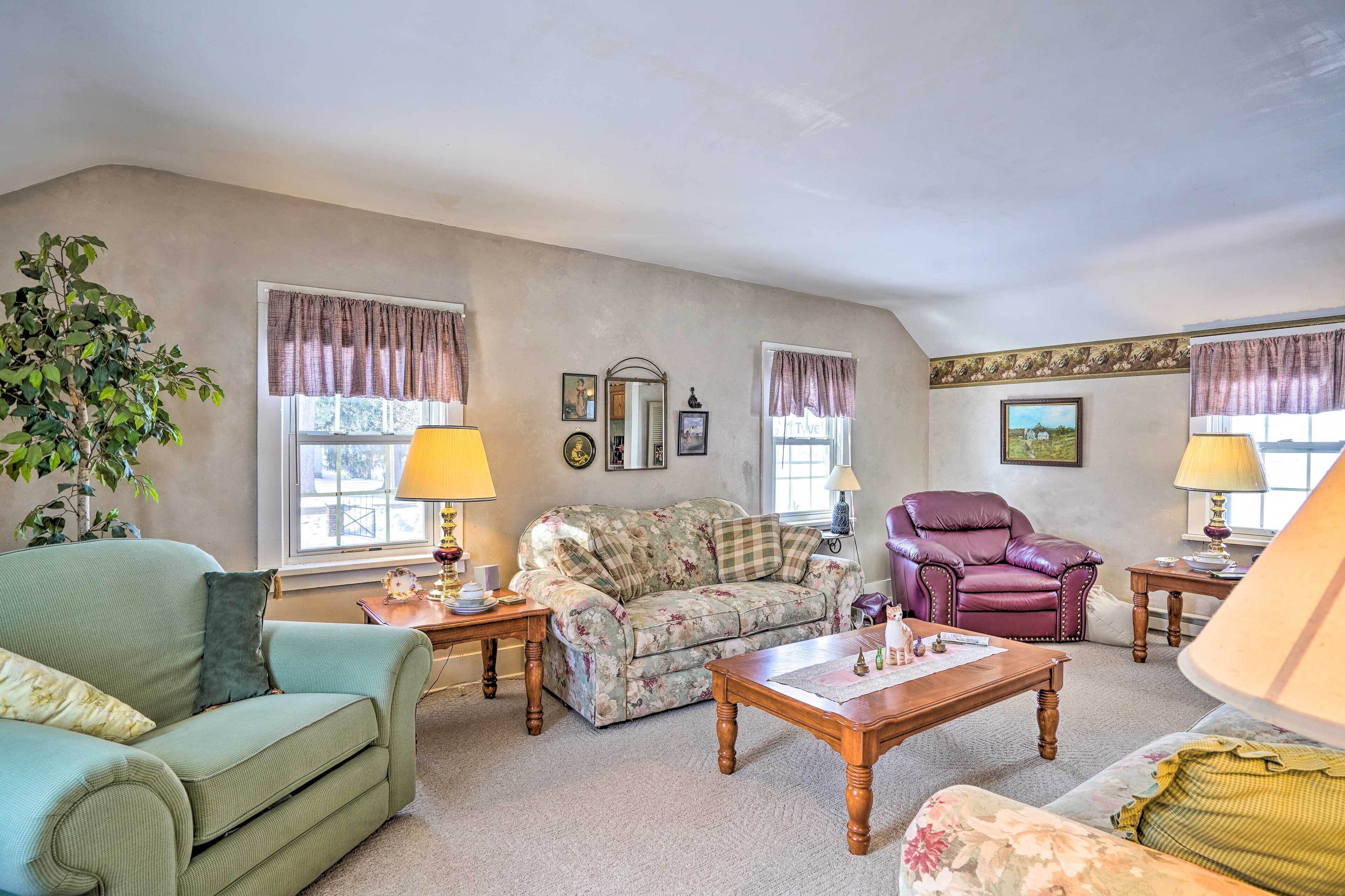 A living room with a green sofa, a floral-patterned sofa, a wooden coffee table, a purple armchair, and a lamp on a side table.