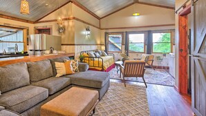 A spacious living area with a large sectional sofa, a small table, and a chair. There is a bed with a metal frame and a window with curtains. The room has wooden floors and a ceiling fan. - Pet-friendly Outdoor Paradise w/ Grill, Decks (Tabor)