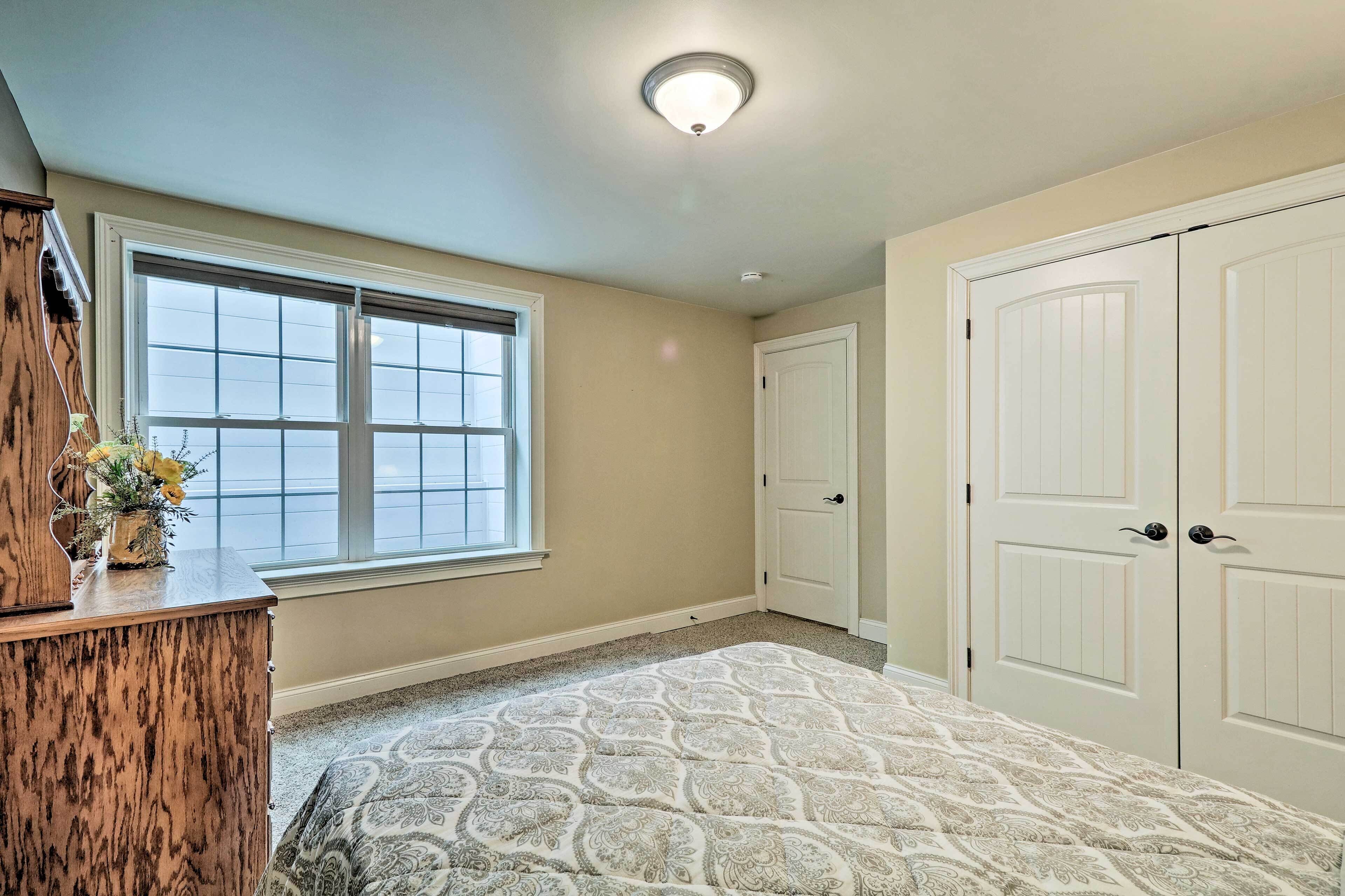 A bedroom with a large window, a wooden dresser, a patterned rug, and a closet with two doors.