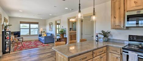 A spacious kitchen with wooden cabinets, a granite countertop, and stainless steel appliances. There is a dining area with a red rug and a blue sofa.