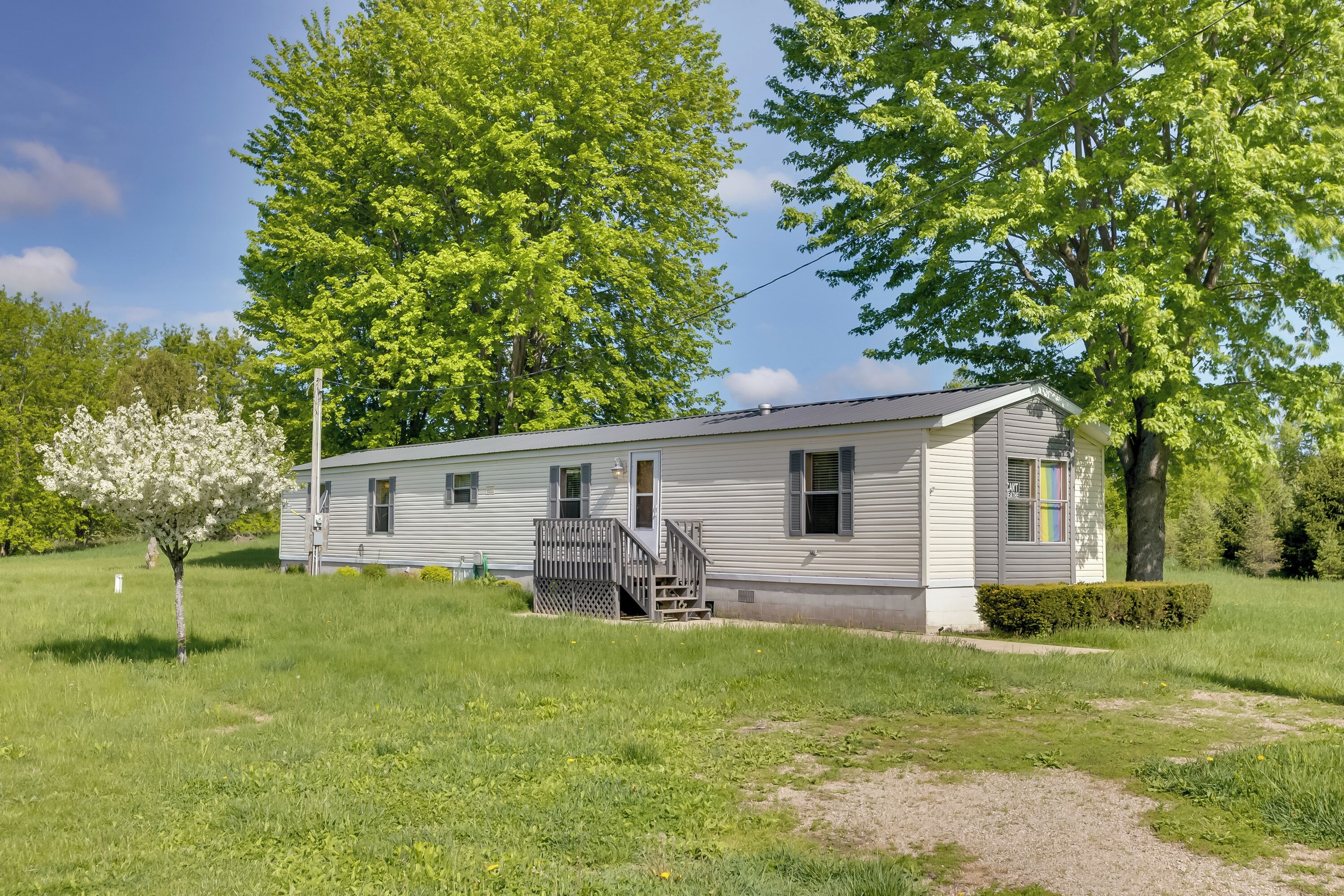 A single-story mobile home with a front porch and a small deck, surrounded by lush green grass and trees.