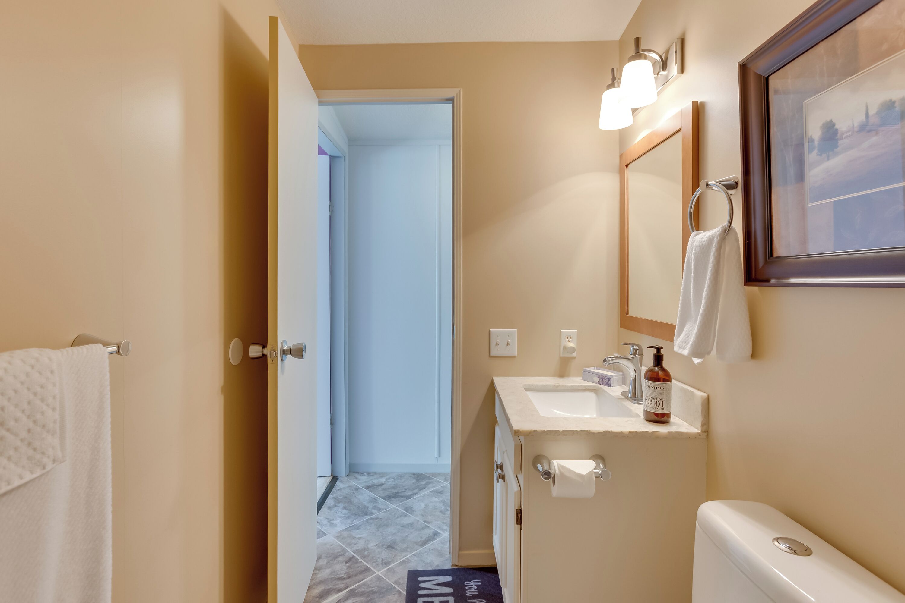 A bathroom with a toilet, sink, and mirror. There is a towel hanging on the wall and a door leading to another room.