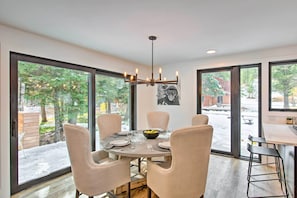 Uma sala de jantar com uma mesa redonda, seis cadeiras, um candelabro e grandes janelas com vista para uma área externa coberta de neve.