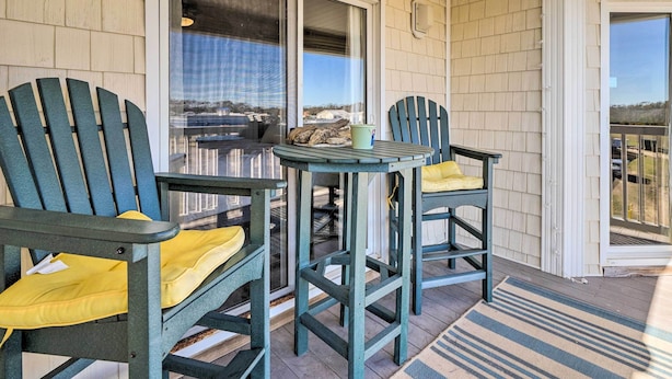 A covered porch with two Adirondack chairs and a small table, overlooking a scenic view. - Oak Island Condo - Walk to the Beach! (Oak Island)