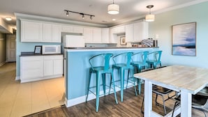 A modern kitchen with white cabinets, a central island with bar stools, and a dining table. - Oak Island Condo - Walk to the Beach! (Oak Island)