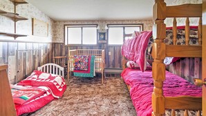 Una habitación rústica con una cama nido de madera, una cama individual con colcha roja, una silla de madera y una cuna con una colcha colorida.