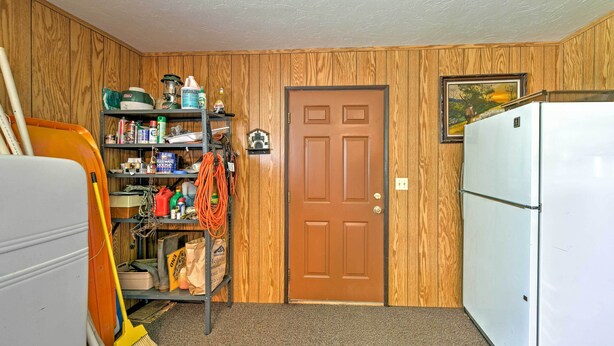 Un cuarto de servicio compacto con paneles de madera, una escoba, un trapeador, un refrigerador blanco y un estante con productos de limpieza.