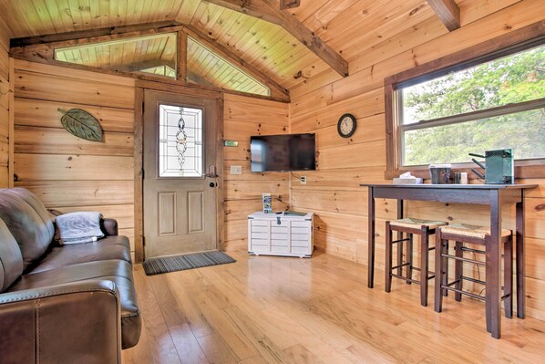 A wooden cabin interior with a sofa, a TV, a small table with stools, and a window with a view of trees. - Hike, Sip & Unwind! Mill Spring Cabin Near Winery (Mill Spring)