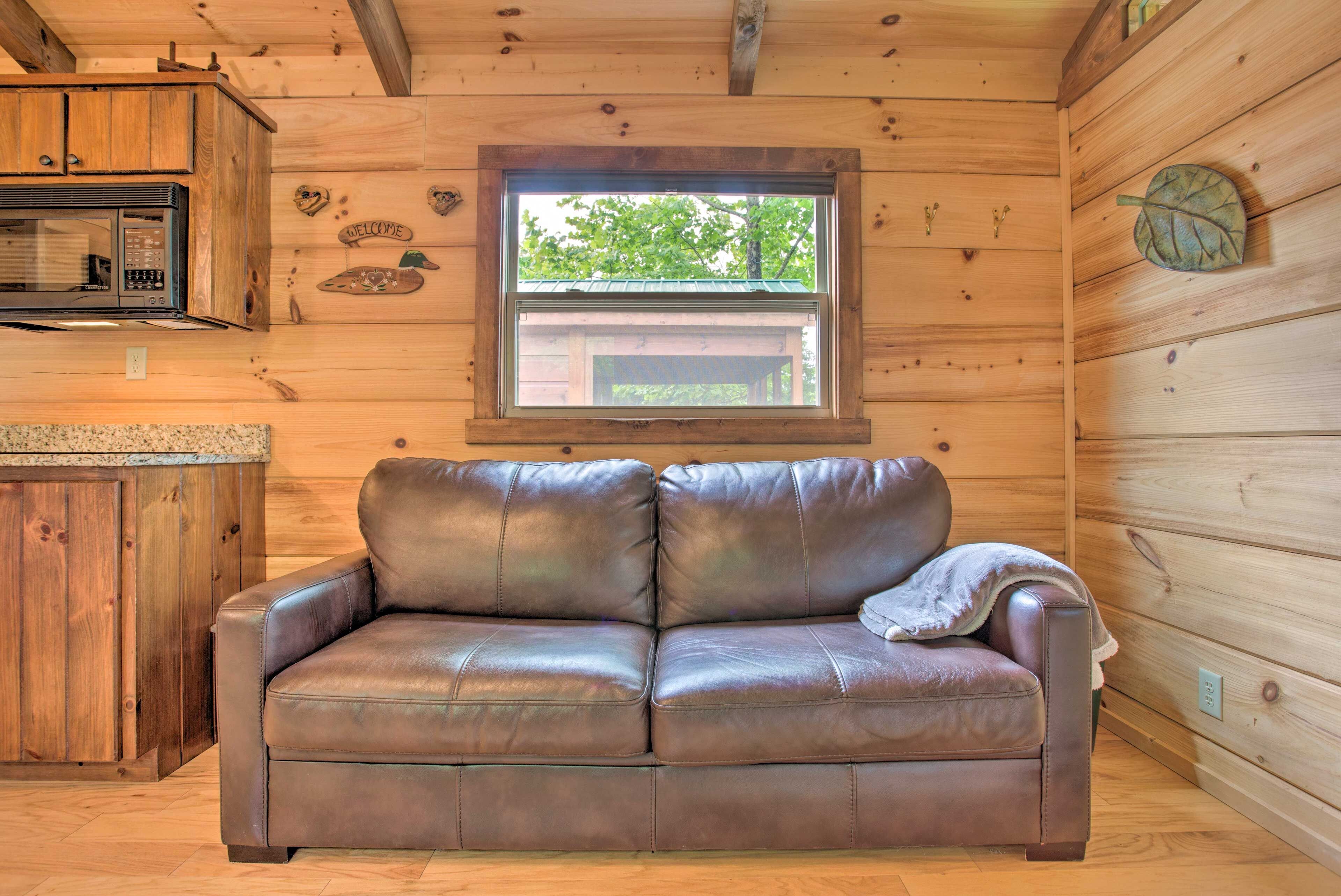 A wooden cabin interior with a leather sofa, wooden cabinets, a microwave, and a window with a view of greenery.