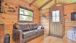 A wooden cabin interior with a leather sofa, a wooden door with a leaf decoration, and a window with a view of trees. - Hike, Sip & Unwind! Mill Spring Cabin Near Winery (Mill Spring)