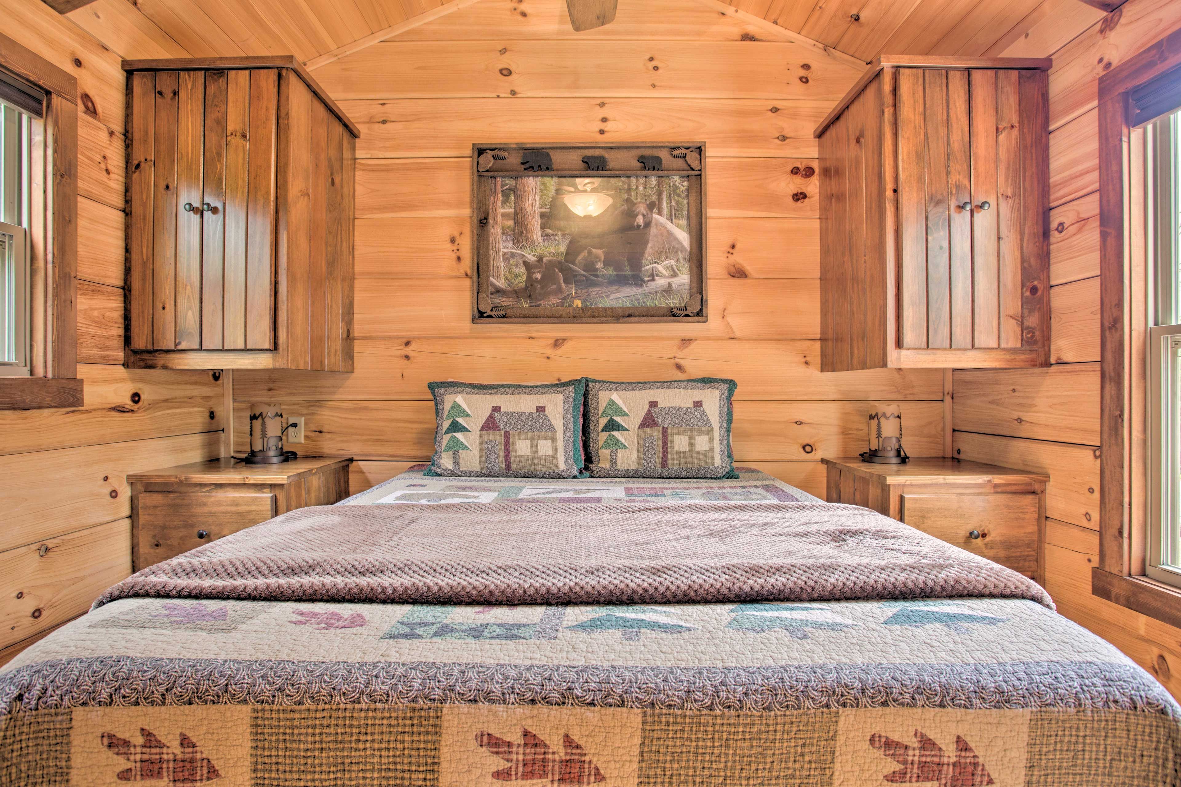 A wooden cabin bedroom with a bed, two nightstands, a framed picture on the wall, and two pillows with a house design.