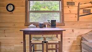 A wooden cabin interior with a wooden table, two woven stools, a window with a view of trees, a clock, and a rustic saw blade hanging on the wall. - Hike, Sip & Unwind! Mill Spring Cabin Near Winery (Mill Spring)