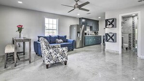 Een ruime woonkamer met een blauwe bank, een gestreept fauteuil, een klein tafeltje met een vaas, en een plafondventilator.