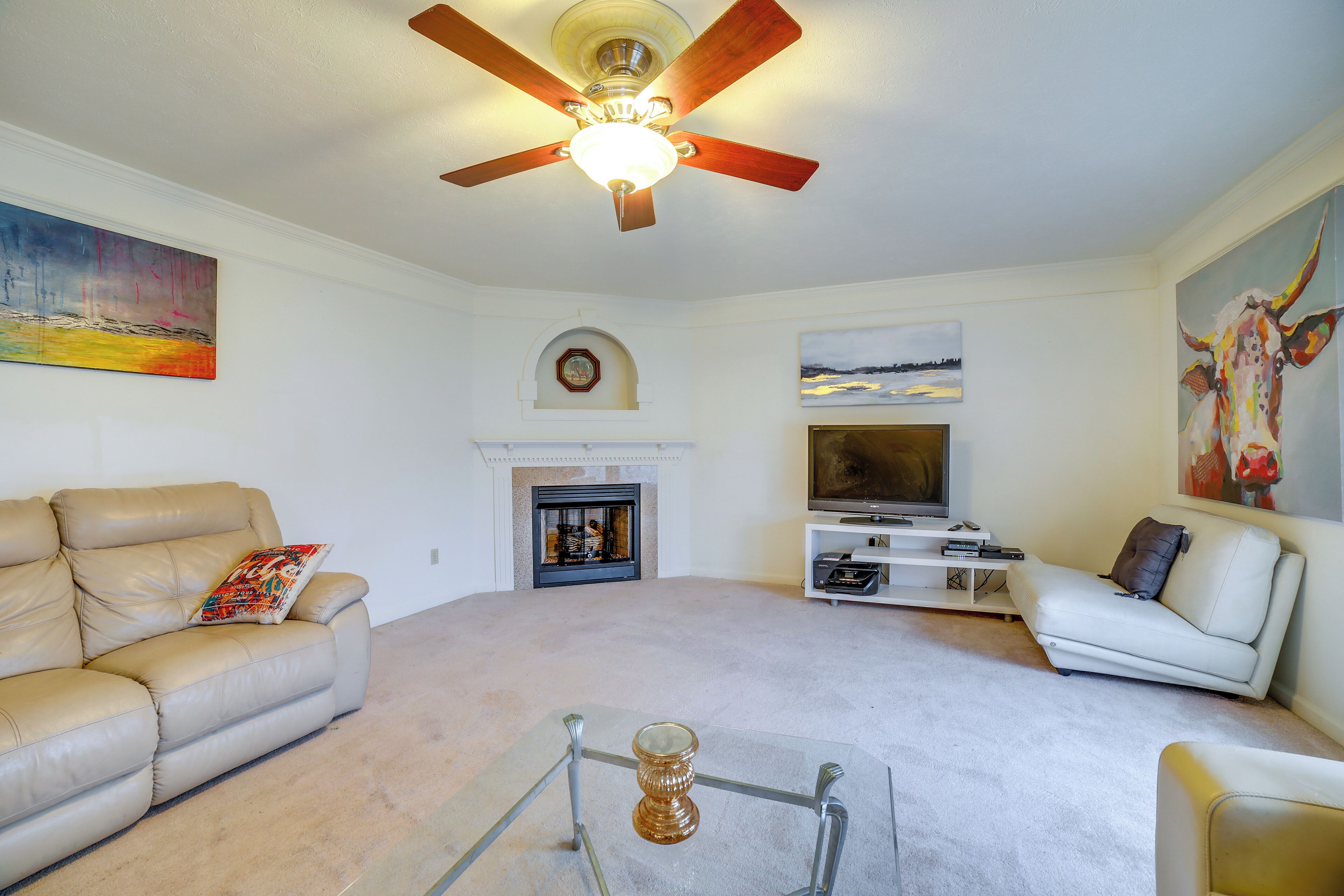 A living room with a beige leather sofa, a white sofa, a glass coffee table, a fireplace, a ceiling fan, and artwork on the walls.