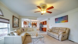 A spacious living room with a glass coffee table, beige sofas, a ceiling fan, and a kitchen in the background. - Pet-friendly Apollo Home w/ Furnished Deck! (Apollo)