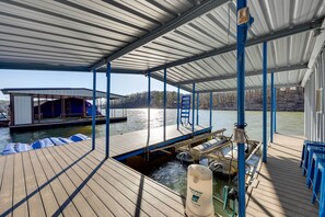 A covered dock with a water pump, a covered seating area, and a building with a blue roof in the background. - Deck, Dock, Fire Pit: Grand Lake Lakefront Home (Jay)