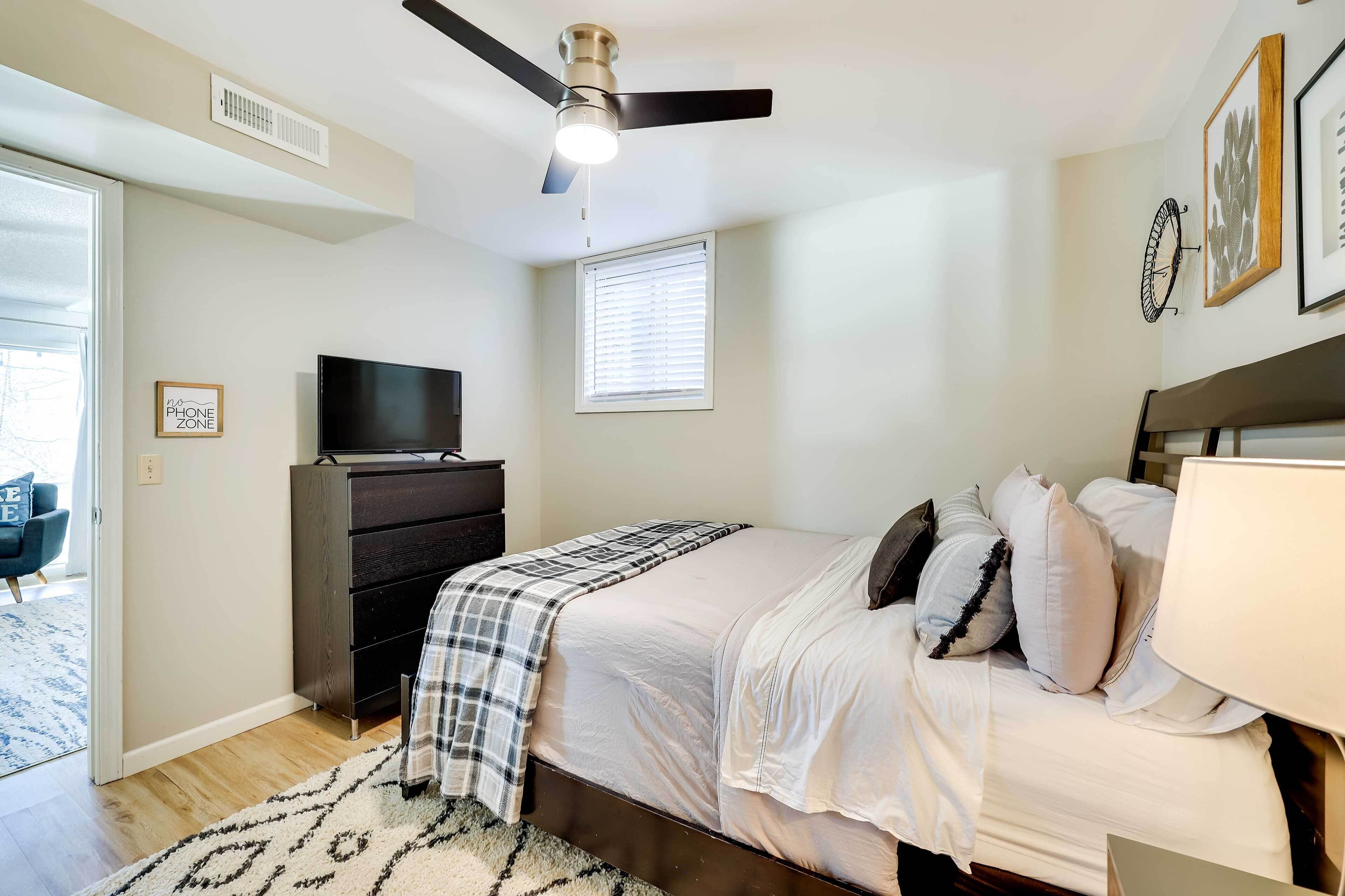 A bedroom with a bed, a television, a ceiling fan, and a window with blinds.