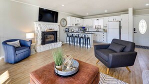 Una sala de estar moderna con chimenea, una cocina con gabinetes blancos y un comedor con taburetes de bar.