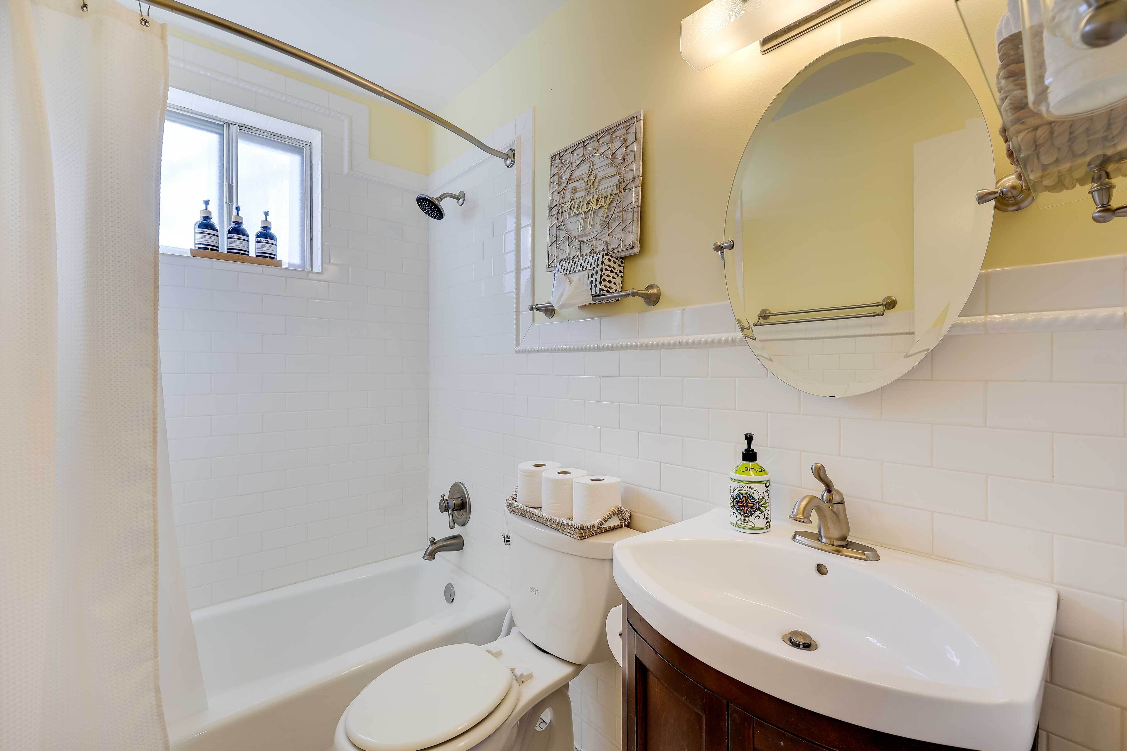 Une salle de bain avec une baignoire, des toilettes et un lavabo. Il y a une fenêtre avec un rideau de douche, un porte-serviettes et un miroir au-dessus du lavabo.