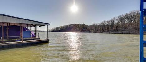 Ein Boot mit überdachtem Deck und einer kleinen Kabine liegt an einem ruhigen Fluss unter einem klaren Himmel, im Hintergrund ein bewaldeter Hügel.