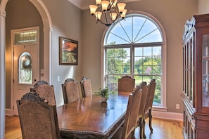 Une salle à manger avec une grande table en bois, six chaises, un lustre, un vitrail et une vue sur la verdure à l’extérieur.