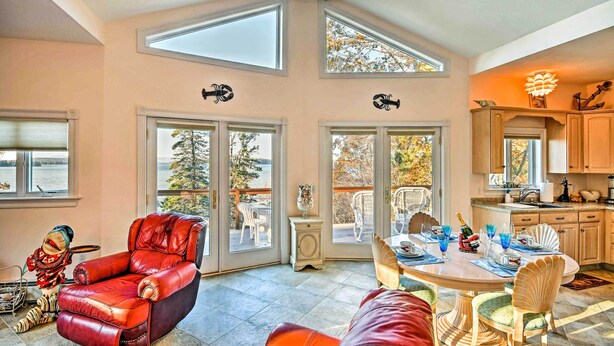 A spacious living room with a red leather chair, a dining table set for four, and a view of the outdoors through large sliding glass doors. - 2BR Coastal Gem! Breathtaking Ocean/mountain View (Sullivan)