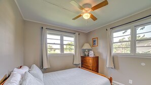 Une chambre à coucher comprenant un lit, un ventilateur de plafond, deux fenêtres avec des rideaux, une table de chevet avec une lampe et une commode.