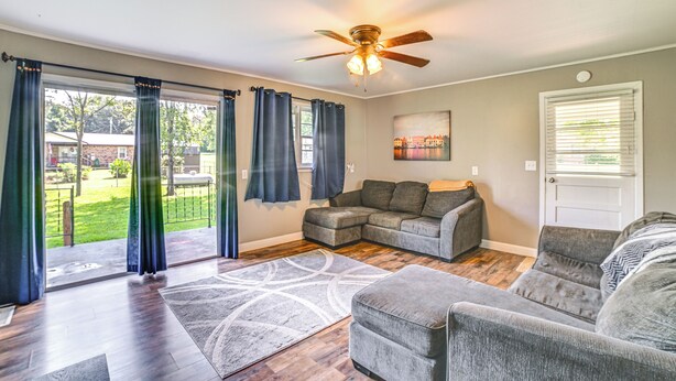 Un salon avec un canapé d’angle, un ventilateur de plafond et une grande fenêtre avec des rideaux.