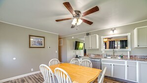 Un coin repas avec une table en bois et des chaises blanches, un ventilateur de plafond, et une cuisine dotée de meubles blancs et d’un plan de travail en granit.