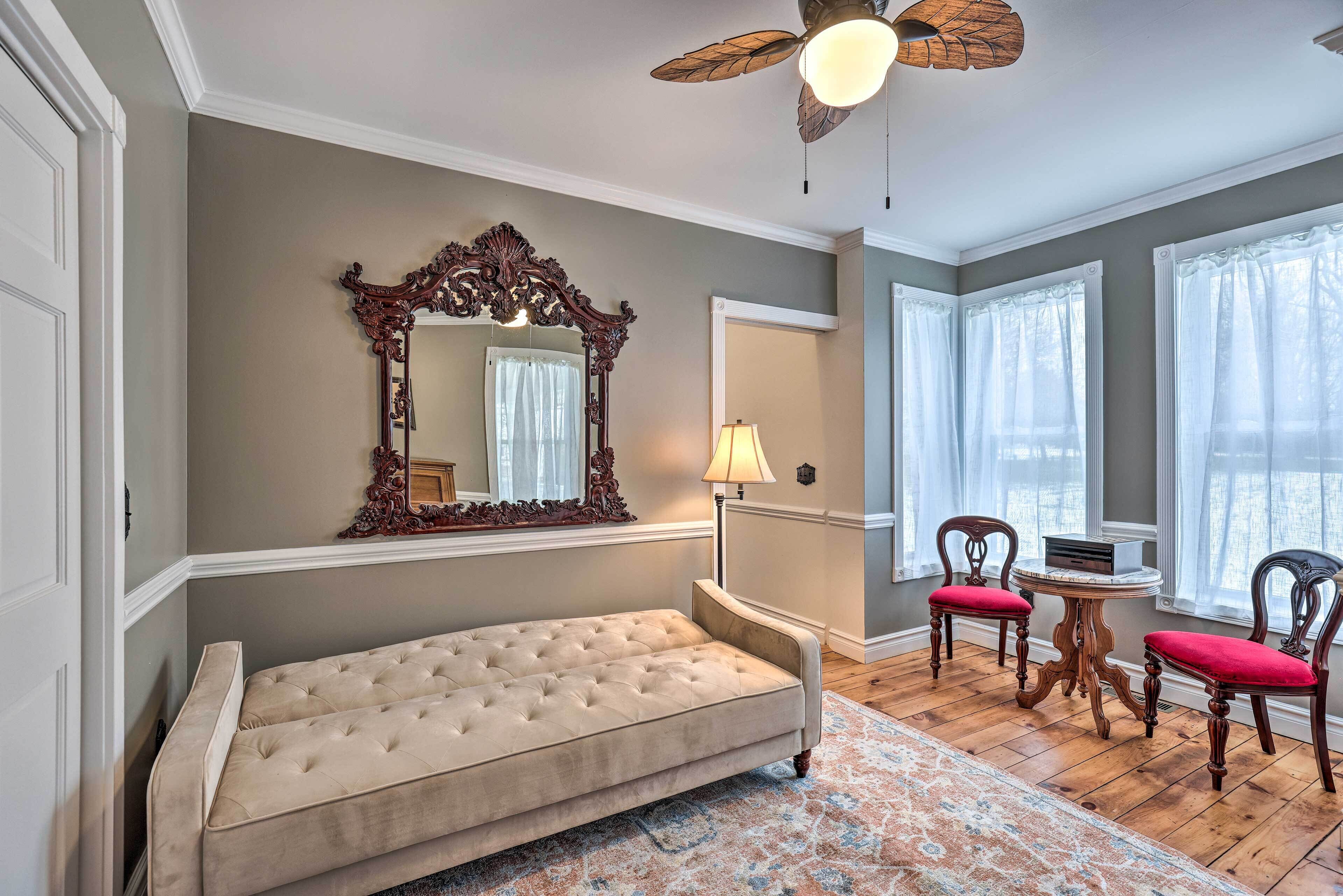 A room with a tufted sofa, a wooden floor, a patterned rug, a wooden chair with red cushions, a table with a lamp, and a large mirror with an ornate frame.