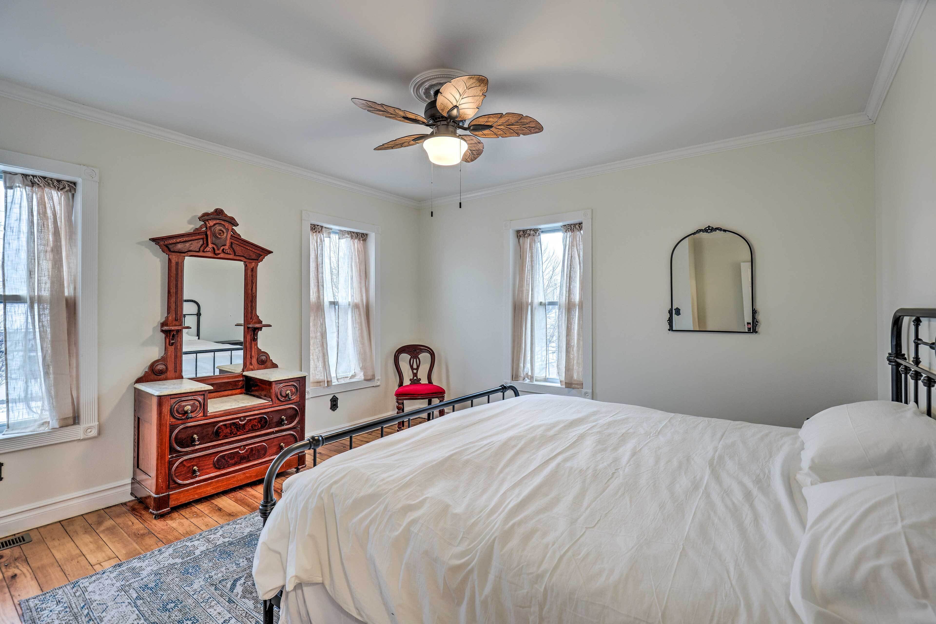A bedroom with a bed, a wooden dresser, a ceiling fan, a mirror, and two windows with curtains.