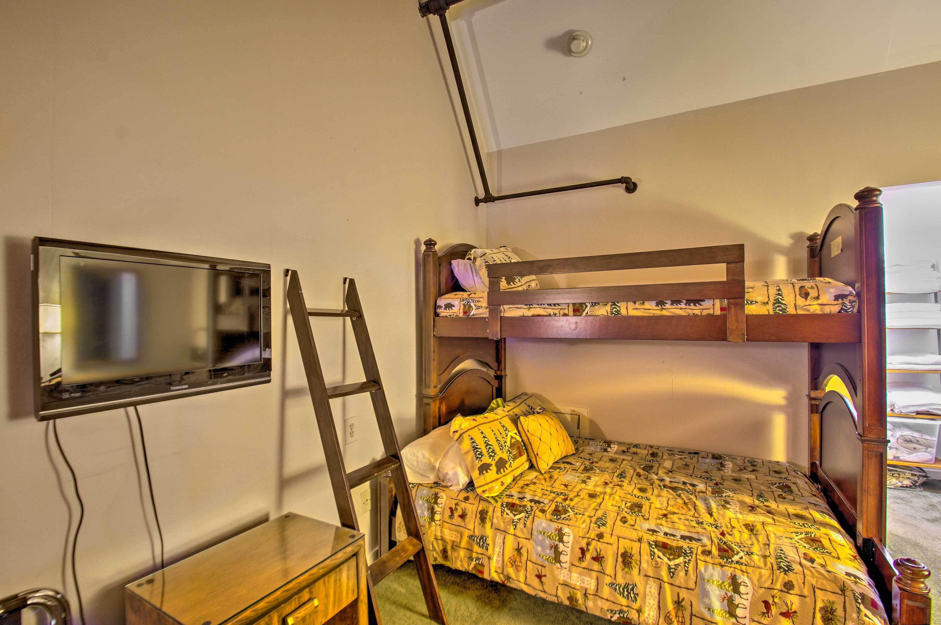 A bunk bed with a patterned bedspread, a wooden desk with a chair, a television mounted on the wall, and a ladder.