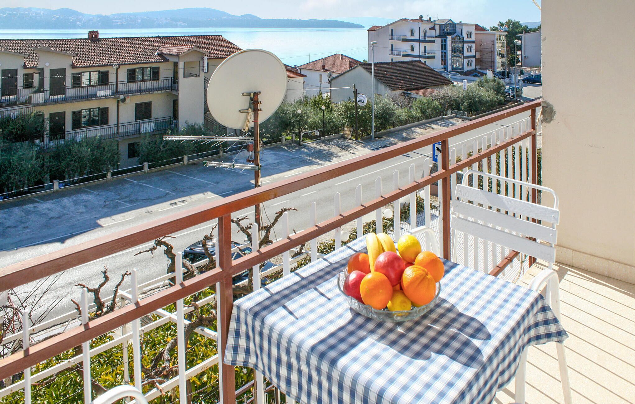 A balcony with a table holding a bowl of fruit, overlooking a street with buildings and a body of water.