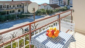 Un balcon avec une table sur laquelle se trouve un saladier rempli de fruits, donnant sur une rue bordée d’immeubles et sur un plan d’eau.