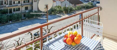 A balcony with a table holding a bowl of fruit, overlooking a street with buildings and a body of water.