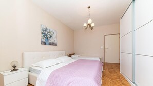 Une chambre à coucher comprenant un lit, des tables de chevet, une commode, un lustre, une porte et un mur orné d’un tableau.