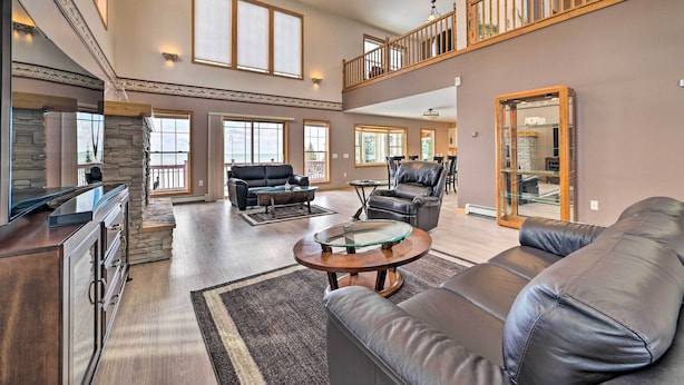 A spacious living room with a high ceiling, a stone fireplace, leather sofas, and a glass coffee table. - Spacious Lakefront Home w/ Sunroom & Bar! (Rogers City)
