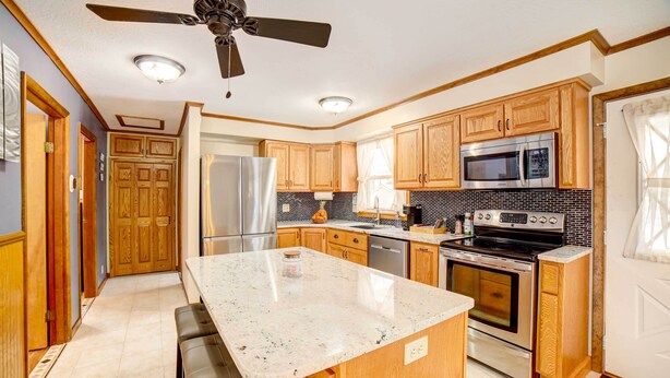 A modern kitchen with wooden cabinets, stainless steel appliances, a large island with seating, and a ceiling fan. - Waterfront Evart House on Chippewa Lake Channel (Evart)
