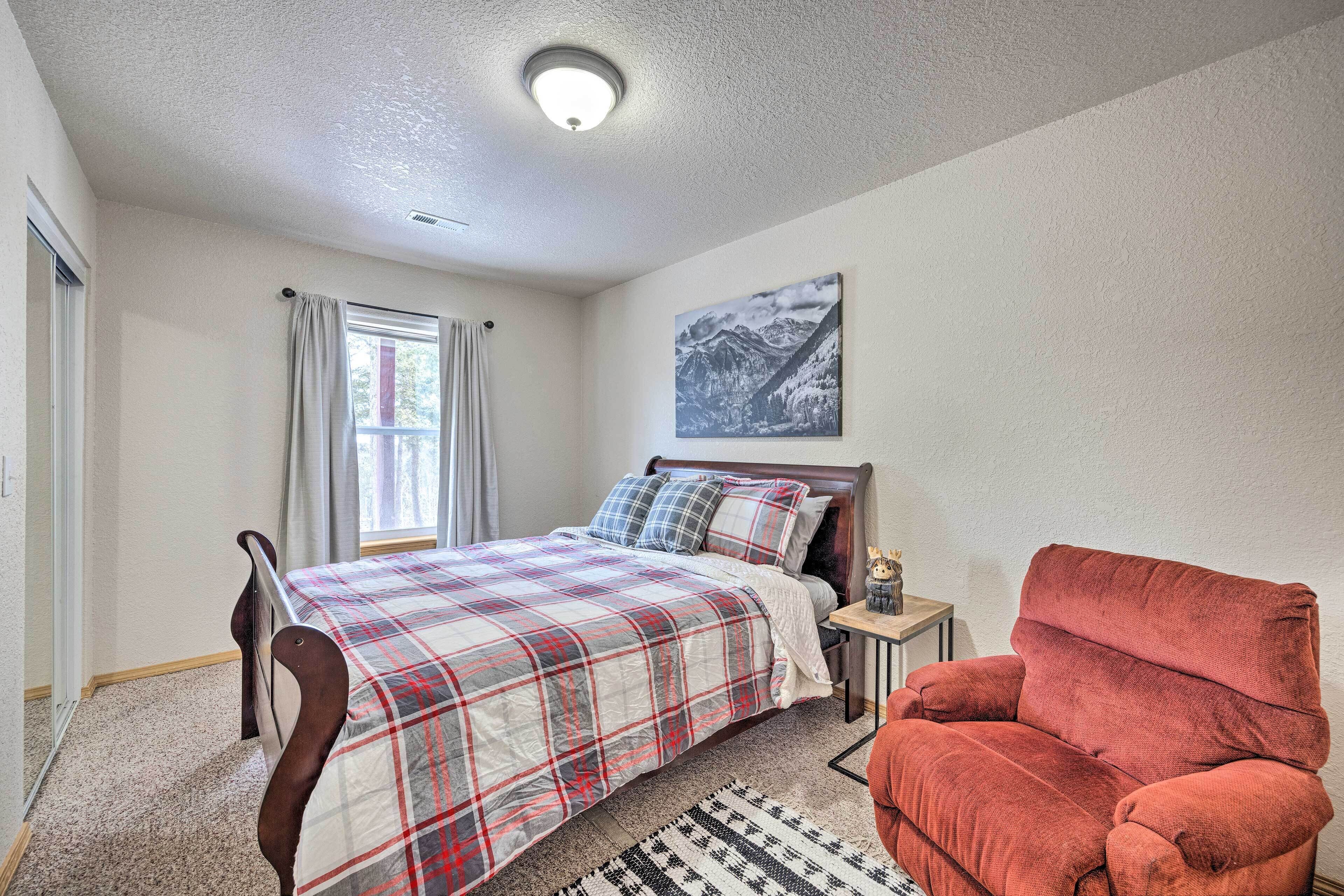 A bedroom with a bed, a red armchair, a nightstand, a window with curtains, and a wall-mounted picture.