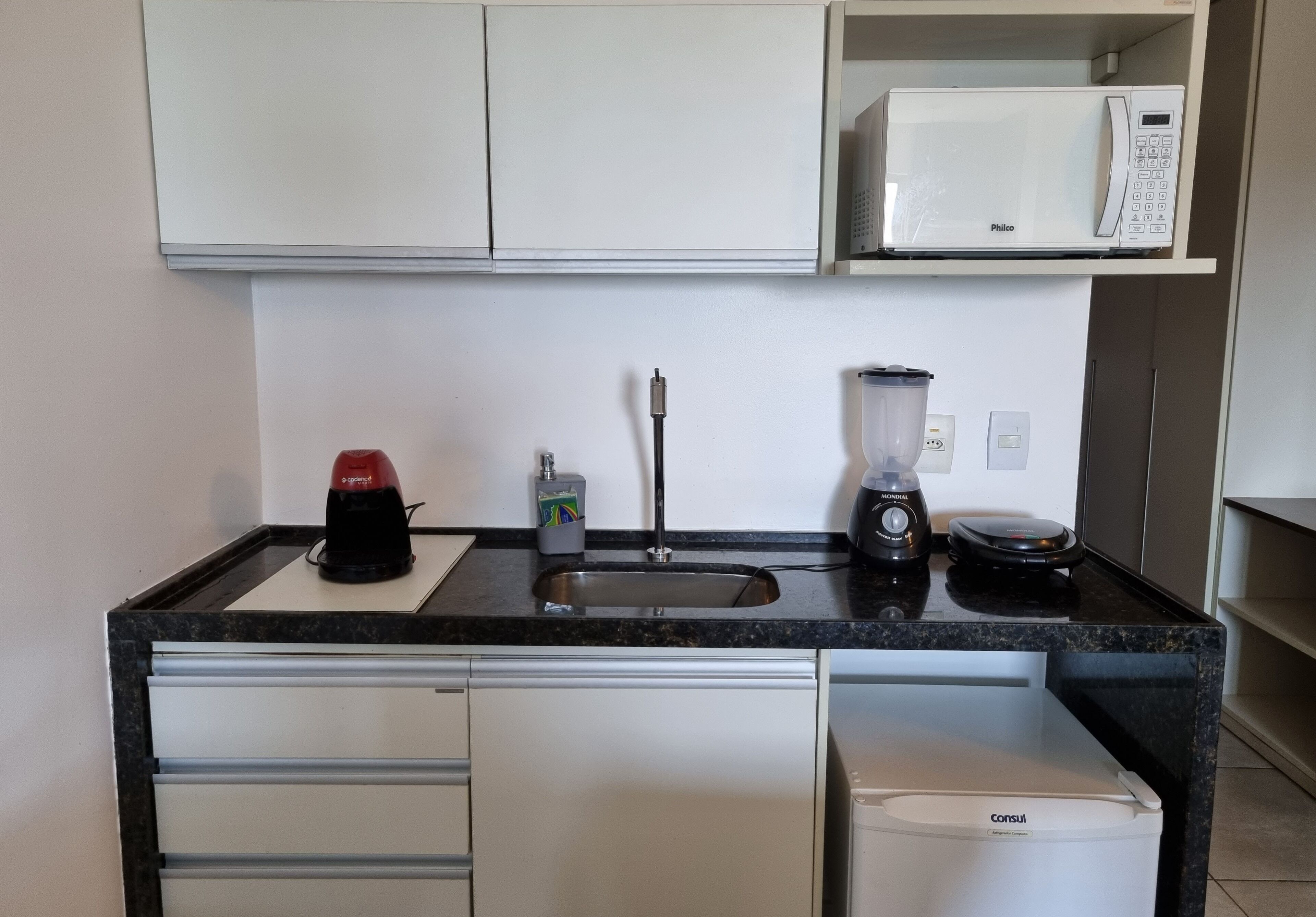 Una cucina compatta con un forno a microonde bianco, una macchina da caffè, un frullatore e un piccolo frigorifero.
