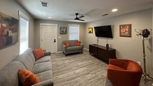 A living room with a grey sofa, orange cushions, a red armchair, a flat-screen TV, a ceiling fan, and artwork on the walls. - Cozy Retro Downtown Living (Albuquerque)